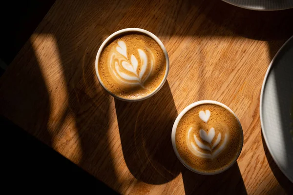 Dua cangkir cappuccino segar dengan seni latte yang indah di atas meja kayu cokelat di bintik-bintik matahari dengan bayangan panjang yang dalam. Delicious kopi pagi dengan susu di kafe. — Stok Foto
