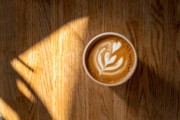 Secangkir kopi dengan seni latte di atas meja kayu. Cappuccino segar dengan latar belakang kayu. Istirahat kopi. Kopi pagi dengan susu. Matahari dan bayangan. Tampilan atas, lay datar, ruang penyalinan — Stok Foto