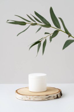 Ahşap kürsüde damgalamak için beyaz plastik kavanoz ve gri arka planda yeşil yapraklı okaliptüs dalı. Doğal güzellik ve spa ürünü sunumu. Boş krema kutusu modeli. Kozmetik reklamcılık