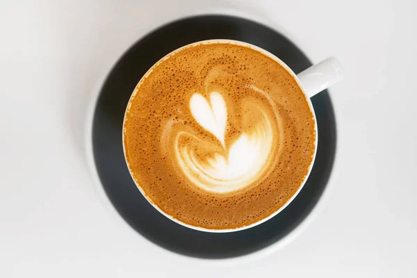 Secangkir kopi segar di piring hitam dekat dengan latar belakang putih. Pagi cappuccino dengan indah desain seni latte hati berbentuk. Tampilan atas — Stok Foto