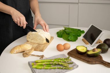Kahvaltı için ekmek kesen mavi önlüklü bir kadın. Sağlıklı yemek için gerekli malzemeler. Tahtada avokado, peçetede kuşkonmaz, yumurta, romantik salata ve tablet. Çevrimiçi tarif