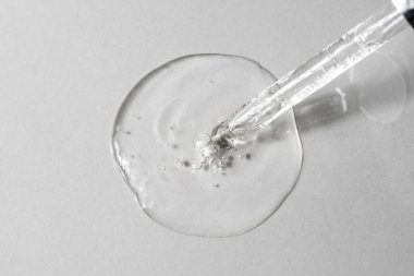 Facial serum with bubbles pouring from glass pipette on grey background. Transparent liquid gel with hyaluronic acid closeup. Anti aging cosmetic product for skin care. Top view, flat lay.