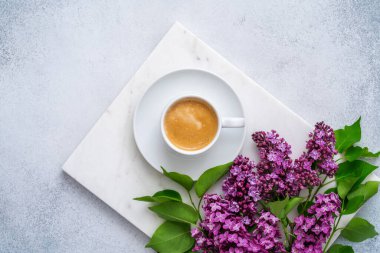Gri arka planda kahve fincanı ve çiçekler. Mermer tepside beyaz kapuçino ve mor leylak. Yeşil yapraklar ve lezzetli içeceklerle açan bahar çiçekleri. Üst manzara, düz uzanma