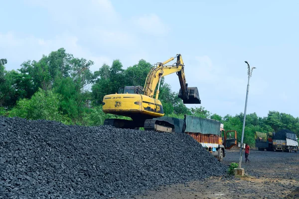 Earth excavator stationed on a heap of charcoal loading coal into a delivery truck with two other trucks standing by. Location: Udipi, Karnataka, India. Date: May 16 2022