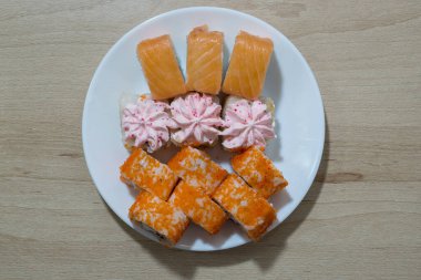 Kırmızı balık, havyar, pirinç ve krema sosundan oluşan üç farklı türdeki rulo ve suşiler beyaz ahşap bir masanın yüzeyinde beyaz bir seramik tabakta düzenlenir..