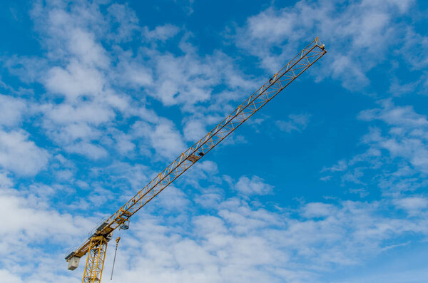The upper part of an industrial construction yellow tower crane with a long boom with stiffeners, concrete counterweight blocks and hooks hanging on chains against a blue sky covered with clouds.