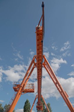 Turuncu üstten kirişli vinç görüntüsü, uzaktan demiryoluna bağlı seyyar taksi, elektrik kalkanları solda yuvarlak köşeli direkler, arka planda bulutlu gökyüzü ve ağaçlar..