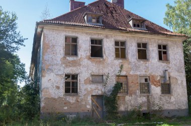 Terk edilmiş eski tuğladan yapılma iki katlı bir apartman dairesi aşınmış alçı kaplı, ahşap çerçevelerden kırılan camlar kısmen çökmüş çatı kiremitleri ve çimenlerle kaplanmış tahta kapılar.