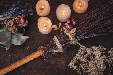 Samhain kutlaması için özel desenli süpürge. El yapımı süpürge, arka planda beyaz mumlar ve renkli kurutulmuş çiçekler ve şifalı otlarla birlikte koyu ahşap bir masada.