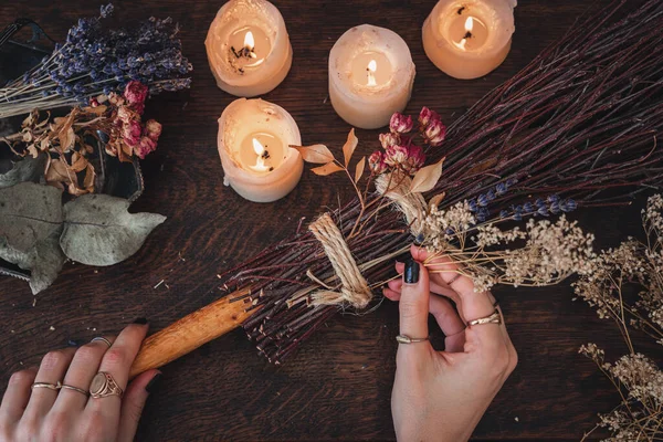 Wiccan女巫为Samhain庆祝会装饰一个Diy专用扫帚 手工制作的扫帚放在黑暗的木制桌子上 背景是点亮的白色蜡烛和五颜六色的干花和香草 — 图库照片