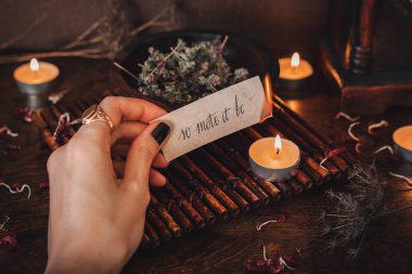 Wiccan witch holding a piece of paper with words So mote it be written on it, and burning it at her altar. Witch casting a spell clipart