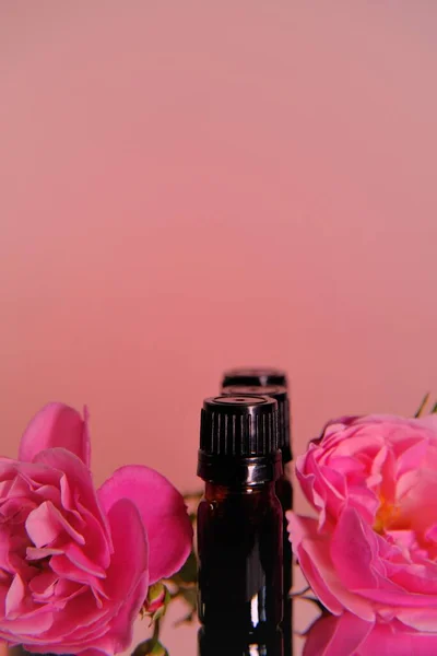 Aceite esencial de rosa.Concepto de aromaterapia y cosmética. Botellas de vidrio marrón y flores de color rosa sobre fondo rosa claro.. — Foto de Stock