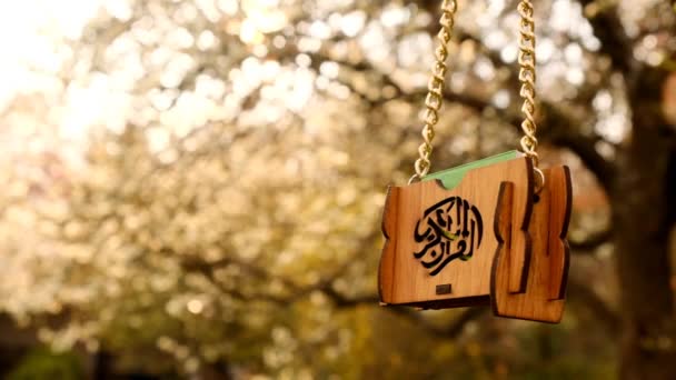 Čas na ramadán. Islámský náboženský symbol. Miniaturní korán v dřevěném pouzdře s islámskou kaligrafií na řetízku na kvetoucích jarních větvích na slunci.Ruka drží mini korán ve větvích s bílými květy — Stock video