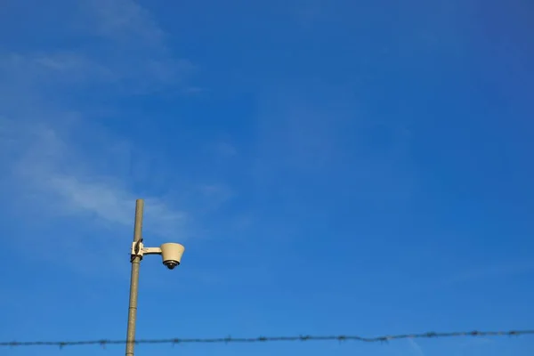 Biztonsági kamera.Videomegfigyelés.Fényképezőgép a szögesdrót mögött, kék ég háttér. Zárt terület. Rendszer objektum — Stock Fotó