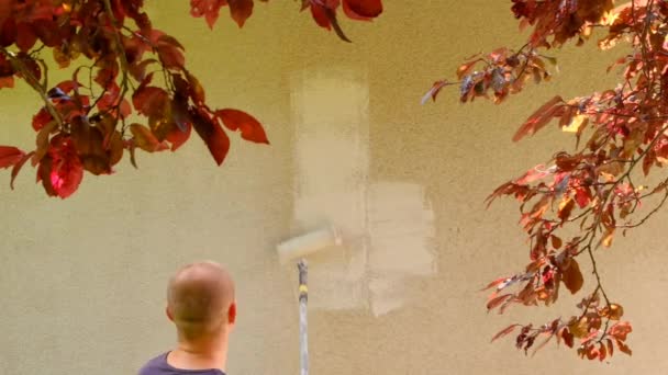 Domácí renovace venku.Muž maluje stěnu doma s válečkem na slunném letním dnu.Nástěnné malby. muž maluje stěny domu v béžové s roller.Repair práce doma — Stock video