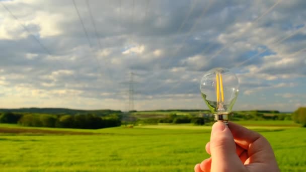 Elektriciteit en groene energie.Lamp in de hand op de Pylon van de elektriciteitsleiding.elektriciteitsleiding. Hoge spanning. Stroomlijnen op blauwe lucht en groene veld achtergrond — Stockvideo