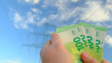 Elektrik ve enerji masrafları. Yeşil enerji. Elektrik fiyatları. Mavi gökyüzünde 100 euro banknot ve elektrik hatları ve yeşil alan arka planı. Para ellerinde. 