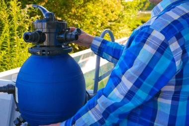 Havuz filtresi. Havuz temizleme ekipmanları. Mavi havuz arka planında ekose gömlekli bir adamın ellerinde mavi su filtresi. Bir adam hortumları havuzun filtresine taşıyor..