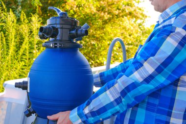 Yüzme havuzu filtresi, havuz temizleme ekipmanları. Mavi su filtresi bir adamın elinde. Bir adam hortumları havuzun filtresine taşıyor..