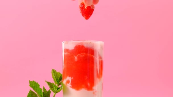 Aardbeiencocktail.hand doet aardbeien in een cocktailglas.Roze cocktail en room in een transparante glazen beker met aardbeien en takjes munt op een roze achtergrond.Berry smoothies. — Stockvideo
