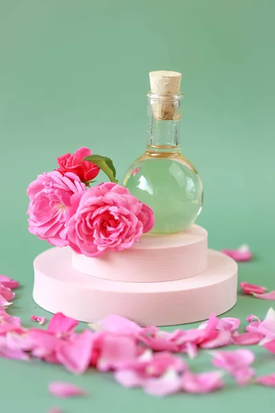 緑の背景にピンクの表彰台にバラの花びらでバラの花のガラス瓶にエッセンシャルオイルをバラ。天然バラ油有機化粧品 — ストック写真