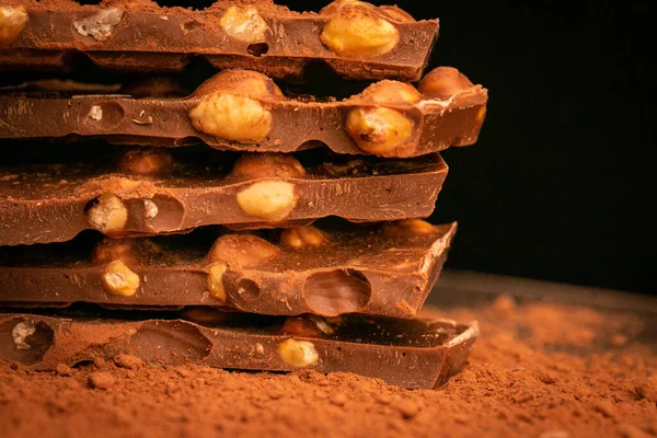 Schokoladenstücke mit Nüssen stapeln sich auf schwarzem Hintergrund. Gebrochene Schokoladentextur. Schokolade mit Haselnüssen und Kakaopulver. Desserts und Süßigkeiten. Vollmilchschokolade mit Nüssen — Stockfoto