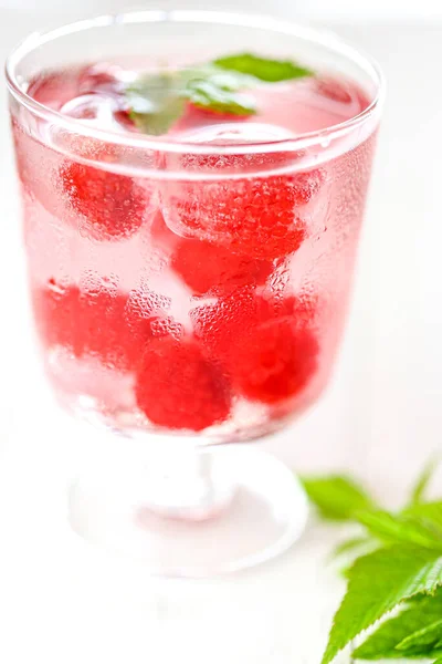 Cocktail di lampone.Bevanda estiva.Bevanda rosa lampone.Cubetti di ghiaccio con lamponi in un bicchiere con foglie di lampone d'acqua su un tavolo di legno bianco. cocktail rinfrescante. — Foto Stock