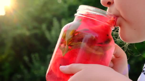 小さな女の子は夏の庭でマグカップからスイカを飲む。子供はガラス製のミストムッグから赤い冷たいカクテルを飲む。スイカの食事スムージー. — ストック動画