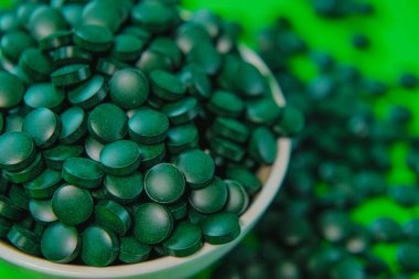 Yuvarlak bir kaseye koyulan bir ilaç. Yeşil arka planda spirulina tabletleri. Vitaminler ve beslenme takviyeleri. Daha besleyici yiyecekler. Sağlıklı bir yaşam tarzı için gıda takviyeleri.