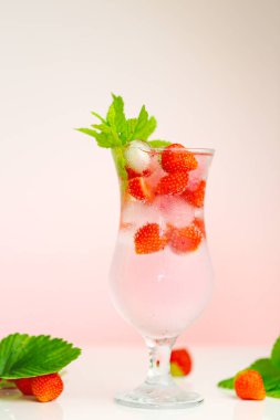 Yaz çileği içeceği. Yaz kokteyli. Buzlu maden suyu bardağı ve açık pembe arka planda yapraklı olgun çilekler. Yaz meyvelerinden iç.