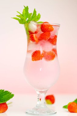 Çilek içkisi. Yaz kokteyllerini tazeleyen içecek. Buzlu maden suyu bardağı ve açık pembe arkaplanlı yapraklı çilek. Yaz meyvelerinden iç.
