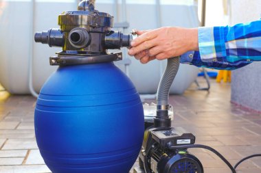  Havuz temizleme ekipmanı. Mavi su filtresi, terasta mavi ekose gömlekli bir adamın elinde. Adam hortumları havuzun filtresine taşıyor..