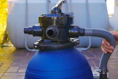  Havuz filtresi. Yüzme havuzu temizleme ekipmanı. Terasta ekose gömlekli bir adamın ellerinde mavi su filtresi. Adam hortumları havuzun filtresine yakın bir yere taşıyor..