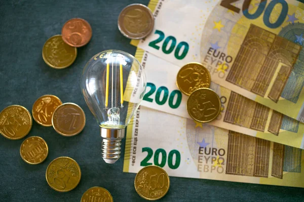 Light bulb, euro banknotes and coins on black chalk board.Increasing the cost of light and heat.Rising electricity prices in Europe.Saving electricity concept.