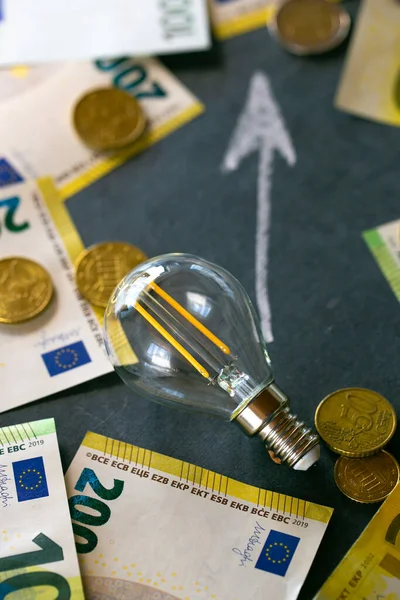 Saving electricity concept.Electricity cost.Rising electricity prices in Europe.Light bulb,euro bills and euro coins, up arrowon on a black chalk board.Crisis of energy production in the EU countries.