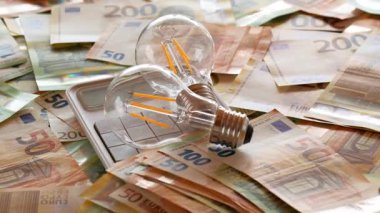 Electricity cost.Two light bulbs on a calculator on euro bills and falling euro cents. Slow motion.Rising electricity prices in Europe.Crisis of energy production in the EU countries.