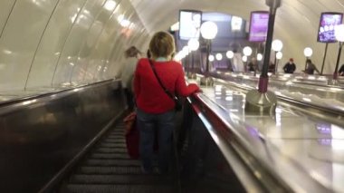 Yürüyen merdivenden metro istasyonuna inen bir kadının arka görüntüsü. Buna paralel olarak, koruyucu maskeli insanlar yürüyen merdivene tırmanırlar. Kapsamlı kısıtlamalar