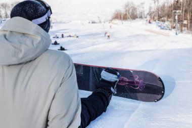 Bir snowboardcu dağın tepesinde oturmuş dinleniyor ve inmeye hazırlanıyor. Arka plan, yüz yok.
