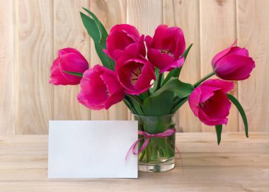 Bouquet pink tulips flowers in glass white vase with white paper card note with space for text on table on a wooden background