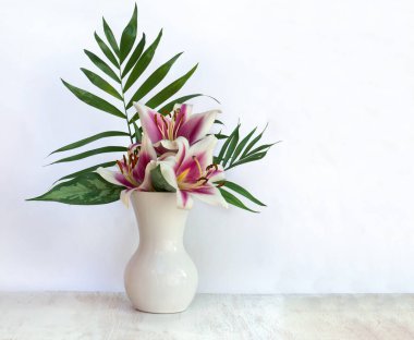 Bouquet pink white flowers lilies with tropical palm leaves and dieffenbachia in porcelain vase on a white wooden table with space for text