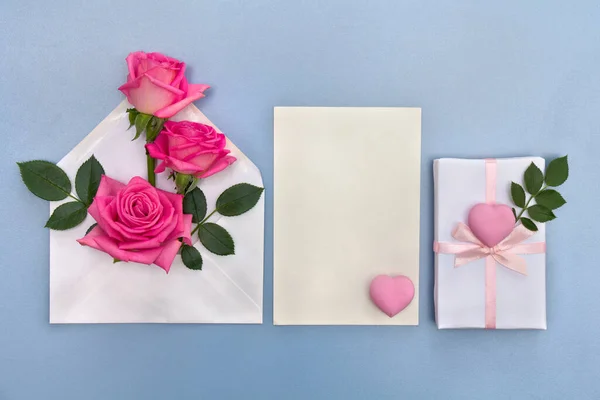 Pink flowers roses postal envelope with paper card note with space for text, pink hearts on a blue background. Top view, flat lay