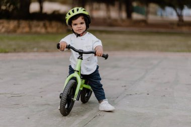 Kasklı küçük çocuk şehir parkında bisiklet sürmeyi öğreniyor. Açık havada neşeli bir çocuk. Kameraya bakıyorum..