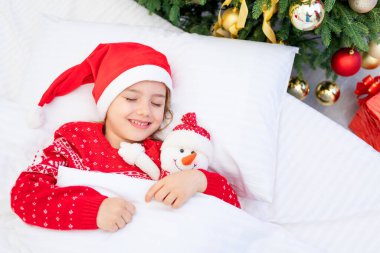 Bir çocuk Noel ağacının altında kırmızı bir süveter ve Noel Baba şapkasıyla uyur. Yeni yıl ya da beyaz bir yatakta kollarında bir kardan adamla.