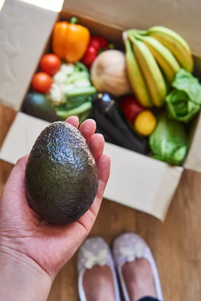 Woman\'s hand holding avocado, box with fruits and vegetables as background. Food delivery concept. Local farm market