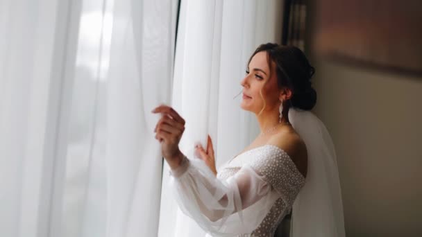 The girl is standing at the window and pushing back the curtain looks at the street. The girl is wearing a white wedding dress