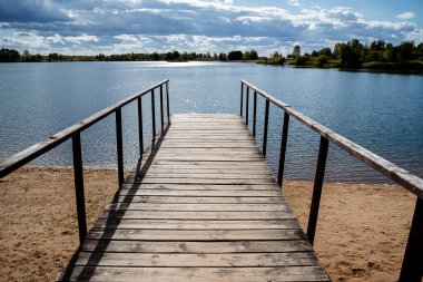 Güneşli bir günde, ormanla çevrili, mavi berrak su kıyısındaki ahşap iskele. Suyun üzerinde güneş parlıyor. Göl kenarında açık hava eğlencesi. Yüksek kalite fotoğraf