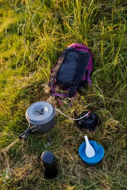 Yürüyüş, açık hava aktiviteleri. Kamp mutfak gereçleri çimenlerin üzerinde: bir termos, bir kupa, bir ısıtıcı, bir kase ve bir sırt çantası. Turist kahvaltısı. Doğada yemek pişirmek. Yüksek kalite fotoğraf