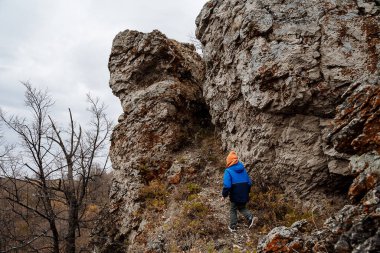 Küçük bir çocuk bir kayaya tırmanarak dağları fetheder. Çocukların aktif turizmi. Dağlarda tek başına yürüyüş. Çocuk sonbaharda doğada sıcacık giyinir. Yüksek kalite fotoğraf