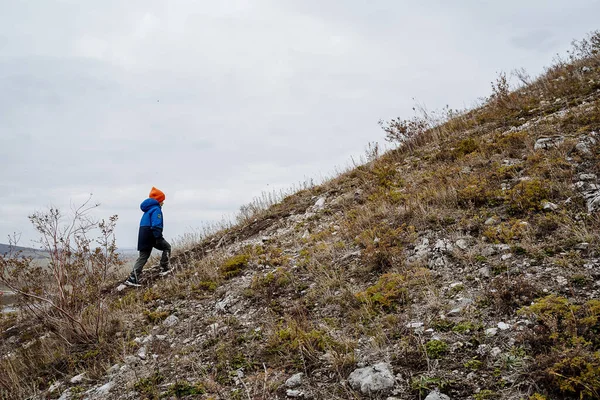 Un niño pequeño con una chaqueta azul sube cuesta arriba. Un joven turista conquista la cima de la montaña. Camina por el camino. Turismo infantil. — Foto de Stock