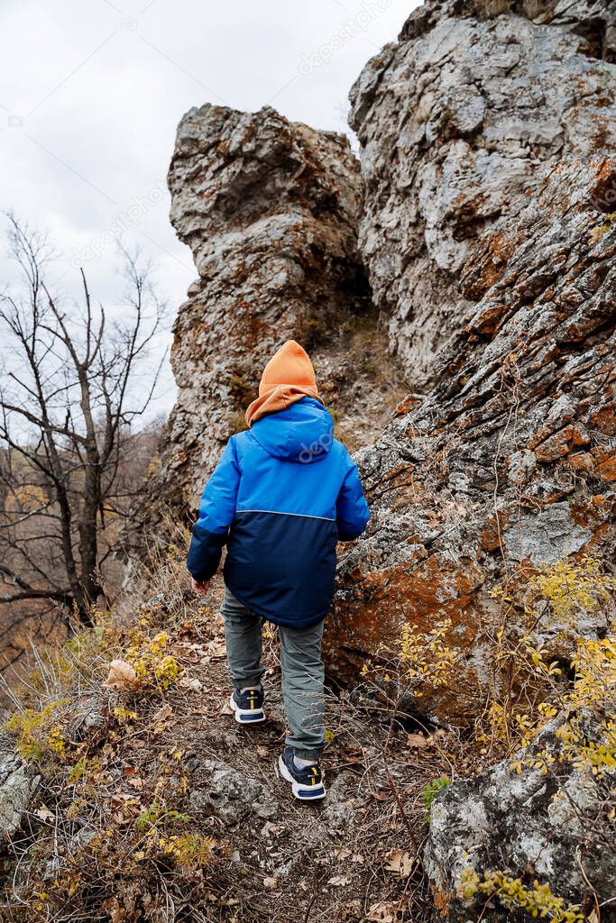 A little boy conquers the mountains climbing a rock. Childrens active tourism. Hiking in the mountains alone. The child is warmly dressed in nature in the fall.
